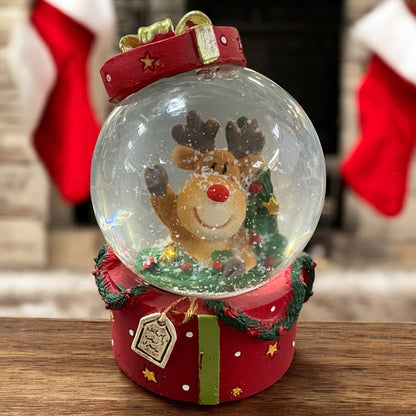 Schneekugel mit Rentierfigur und Tannenzweig, liebevoll in weihnachtlicher Kulisse mit Kaminfeuer inszeniert.