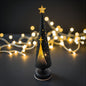Moderne LED-Dekosäule in Tannenbaumform in Schwarz mit filigranen Stern- und Lichtpunkten, stimmungsvoll beleuchtet vor warmem Lichterketten-Hintergrund.