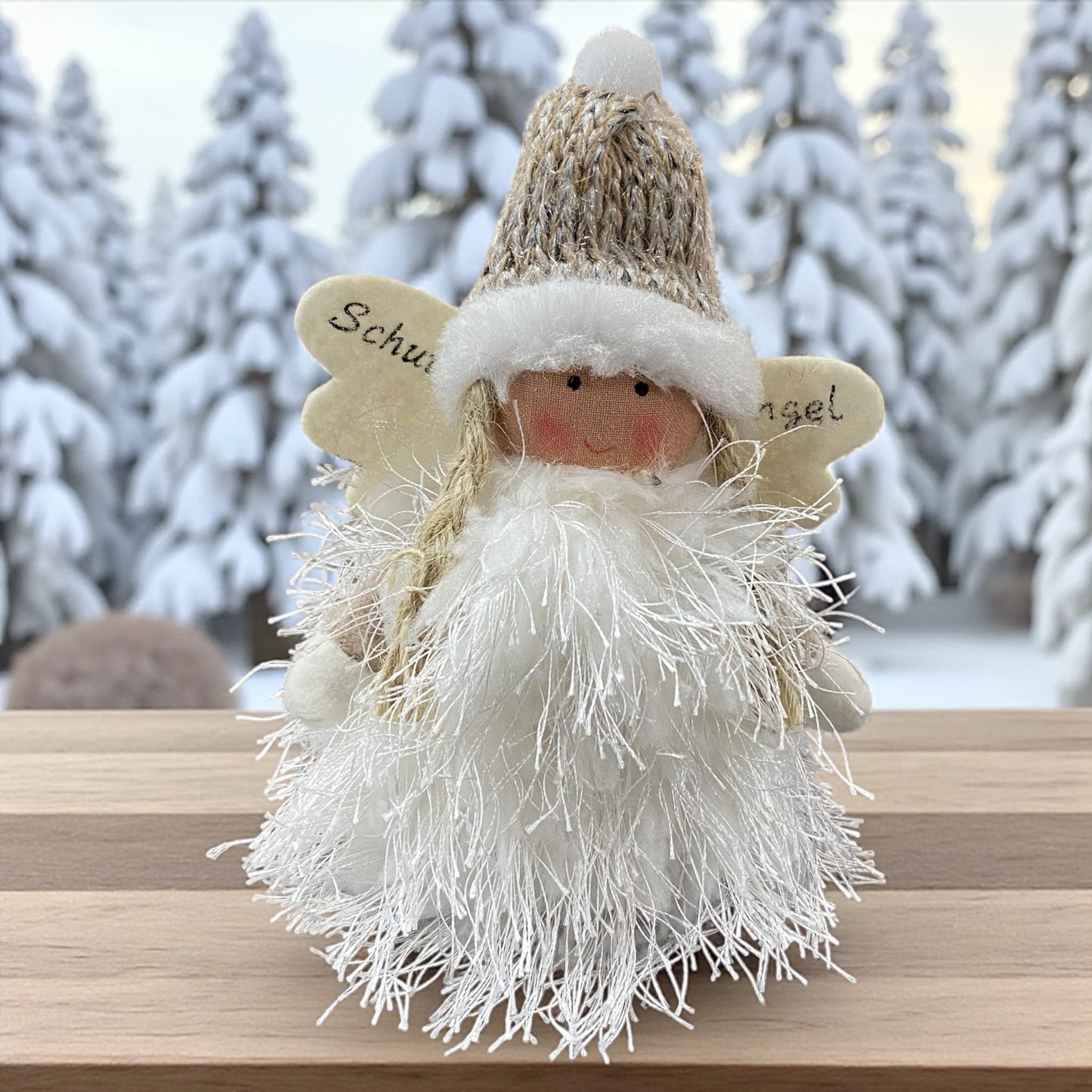 Flauschige Stoff-Engelfigur mit beiger Strickmütze und weißen Flügeln auf einem Holztisch, im Hintergrund verschneite Bäume.