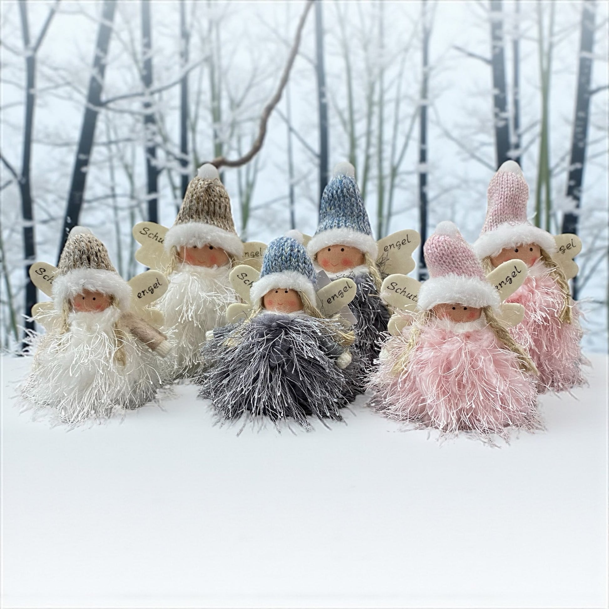 Kleine Stoff-Engelfiguren mit flauschigen Kleidern und Strickmützen in den Farben Weiß, Grau und Rosa, jeweils mit Flügeln und der Aufschrift „Schutzengel“ im Ambiente