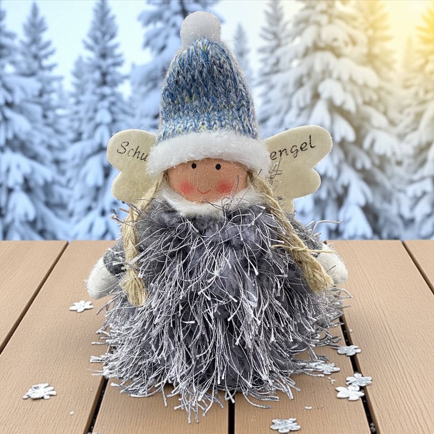 Flauschige Stoff-Engelfigur mit blauer Strickmütze und weißen Flügeln auf einem Holztisch, im Hintergrund verschneite Bäume.