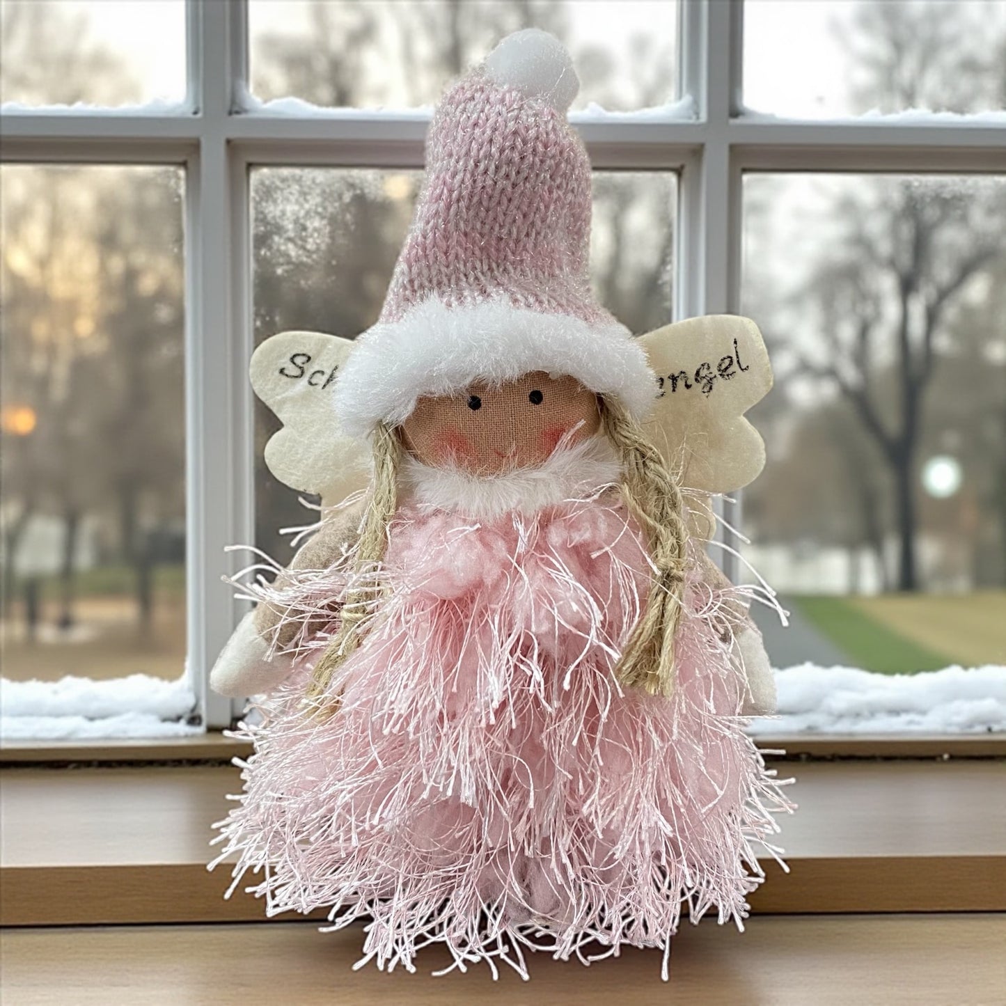 Flauschige Stoff-Engelfigur mit roa Strickmütze und weißen Flügeln auf einem Holztisch, im Hintergrund verschneite Bäume.