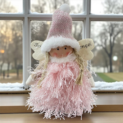 Flauschige Stoff-Engelfigur mit roa Strickmütze und weißen Flügeln auf einem Holztisch, im Hintergrund verschneite Bäume.