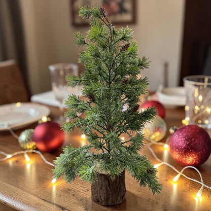 Ein dekorativer Mini-Tannenbaum auf einem Holzsockel steht auf einem festlich gedeckten Tisch.