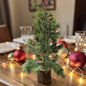 Ein dekorativer Mini-Tannenbaum auf einem Holzsockel steht auf einem festlich gedeckten Tisch.