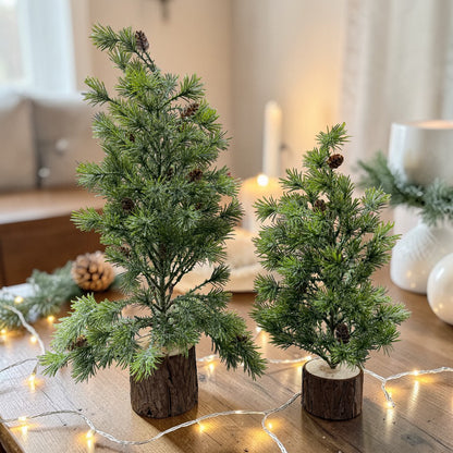 Zwei dekorative Mini-Tannenbäume mit Holzsockel stehen auf einem Tisch mit Lichterkette im Hintergrund.