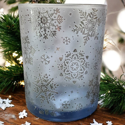 Dekoratives blaues Teelichtglas mit Schneeflocken-Ornamenten, weihnachtlich arrangiert mit Tannenzweigen.