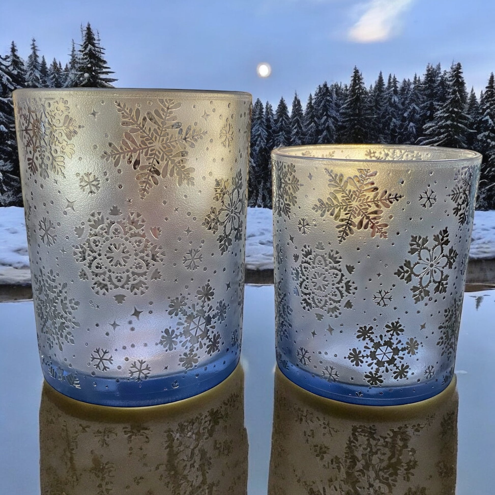 Zwei blaue Teelichtgläser mit filigranen Schneeflockenmustern, stimmungsvoll im winterlichen Ambiente beleuchtet.