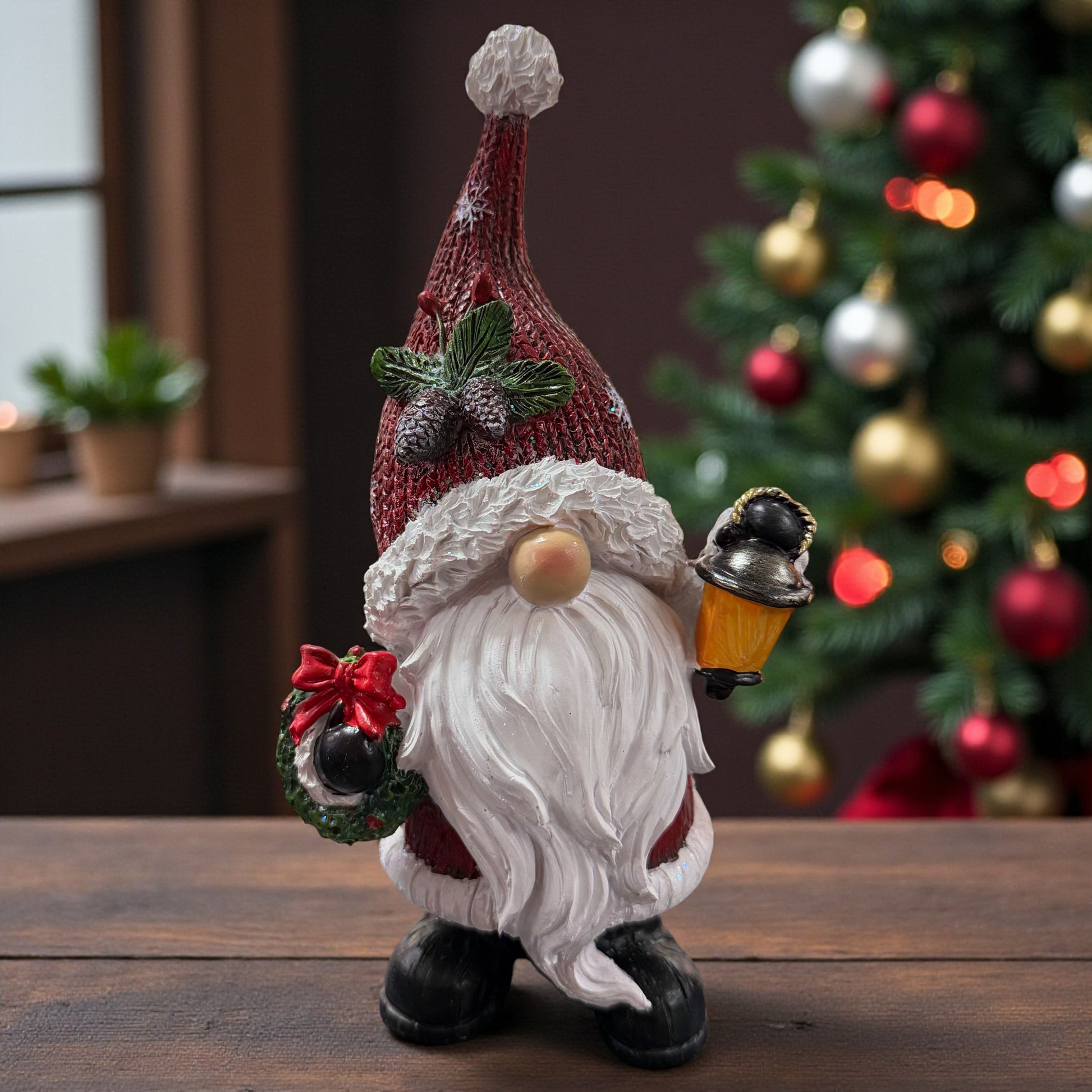 Der Wichtel mit Tannenzapfen-Dekor auf der Mütze, Kranz und Laterne sorgt vor einem geschmückten Weihnachtsbaum für eine stimmungsvolle und gemütliche Atmosphäre.