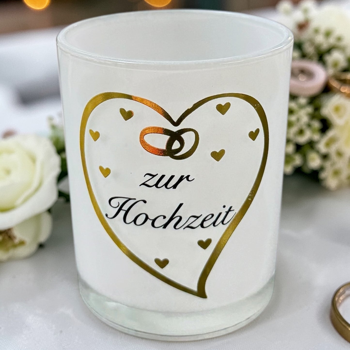 Weißes Glaswindlicht mit goldenem Herzmotiv, kleinen Herzsymbolen und dem Schriftzug „zur Hochzeit“ auf dekoriertem Untergrund mit Blumen im Hintergrund.