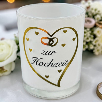 Weißes Glaswindlicht mit goldenem Herzmotiv, kleinen Herzsymbolen und dem Schriftzug „zur Hochzeit“ auf dekoriertem Untergrund mit Blumen im Hintergrund.