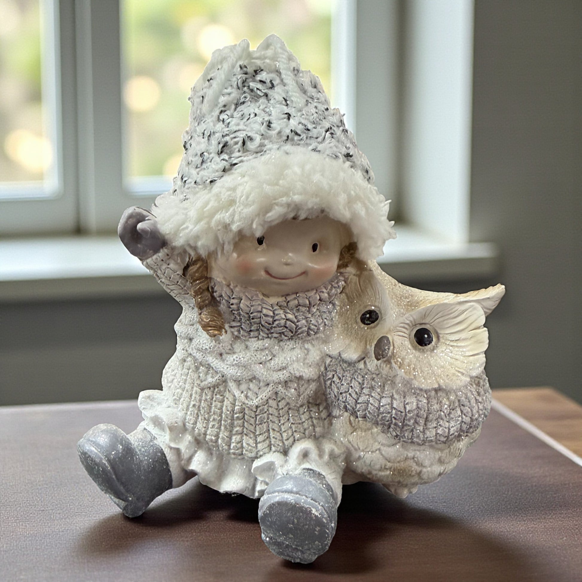 Keramikfigur zeigt ein sitzendes Mädchen mit Winterkleidung und Mütze neben einer Eule auf einer Tischoberfläche vor einem Fenster.