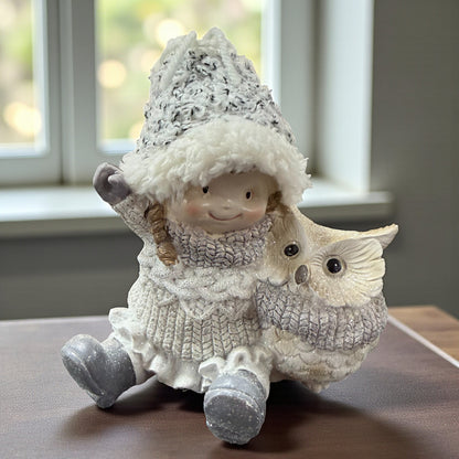 Keramikfigur zeigt ein sitzendes Mädchen mit Winterkleidung und Mütze neben einer Eule auf einer Tischoberfläche vor einem Fenster.