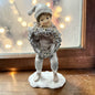 Eine dekorative Figur zeigt ein Kind in winterlicher Kleidung, das einen Kranz hält.