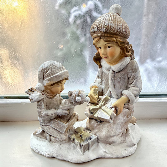 Eine dekorative Figur zeigt zwei Kinder beim Auspacken von Geschenken auf einer winterlichen Basis.