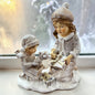 Eine dekorative Figur zeigt zwei Kinder beim Auspacken von Geschenken auf einer winterlichen Basis.
