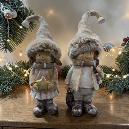 Eine Mädchen- und eine Jungenfigur in winterlicher Kleidung stehen auf einer Holzoberfläche vor einer dekorierten Tannengirlande mit Lichterkette.