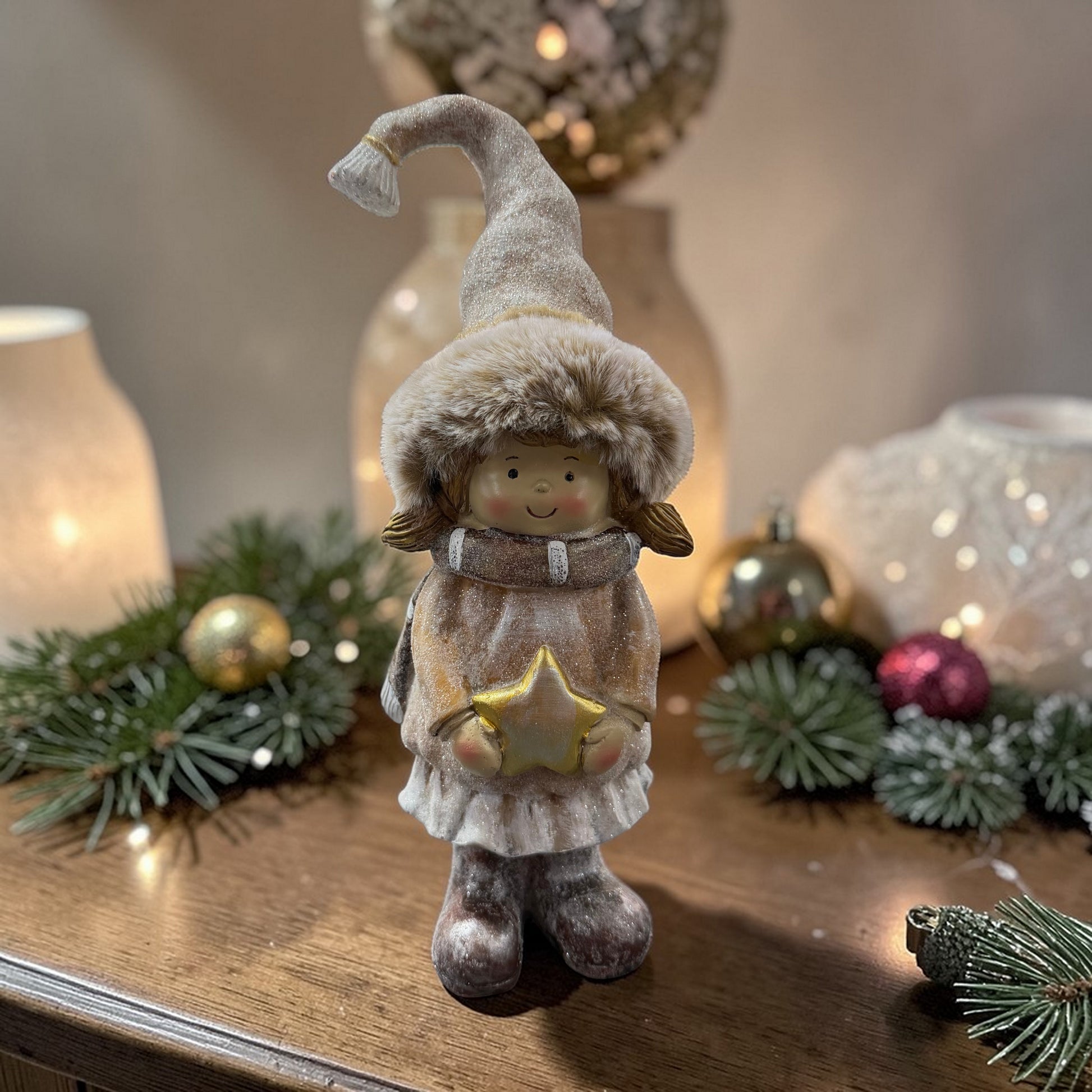 Eine dekorative Jungenfigur mit Fellmütze und Schal steht auf einer Holzoberfläche vor einer Lichterkette und winterlicher Dekoration.
