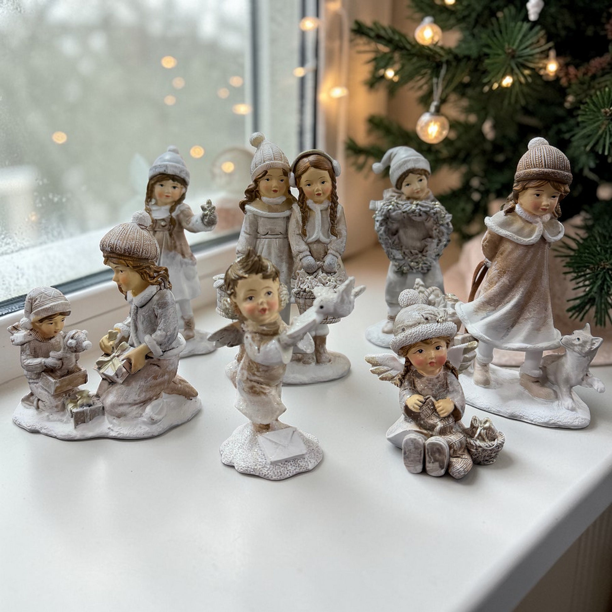 Mehrere dekorative Figuren mit Kindern in winterlicher Kleidung sind auf einer Fensterbank neben einem Weihnachtsbaum arrangiert.