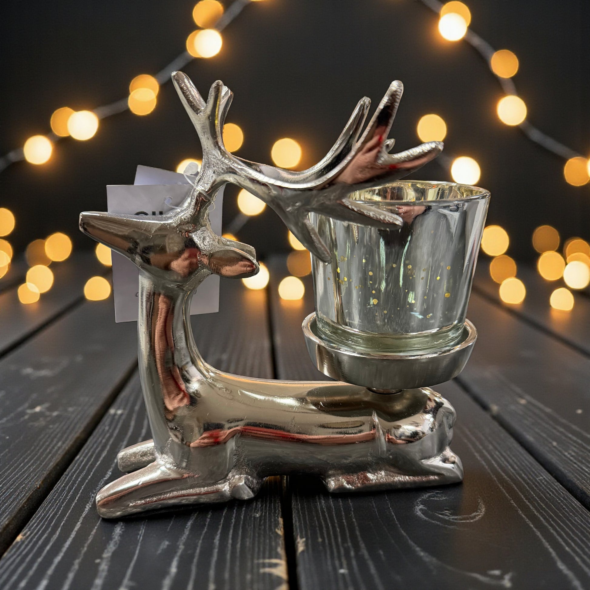 Dekorativer Teelichthalter in Form eines Hirsches aus glänzend silberfarbenem Aluminium mit eingesetztem Glas-Windlicht, arrangiert auf dunkler Holzoberfläche vor warm leuchtender Lichterkette im Hintergrund.