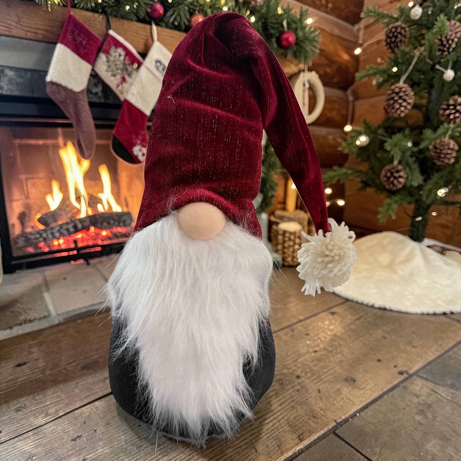 Wichtelfigur aus Stoff mit schwarzem Körper, weißem Bart und roter Mütze auf einem Holzboden vor einem Kamin mit Weihnachtsdekoration.