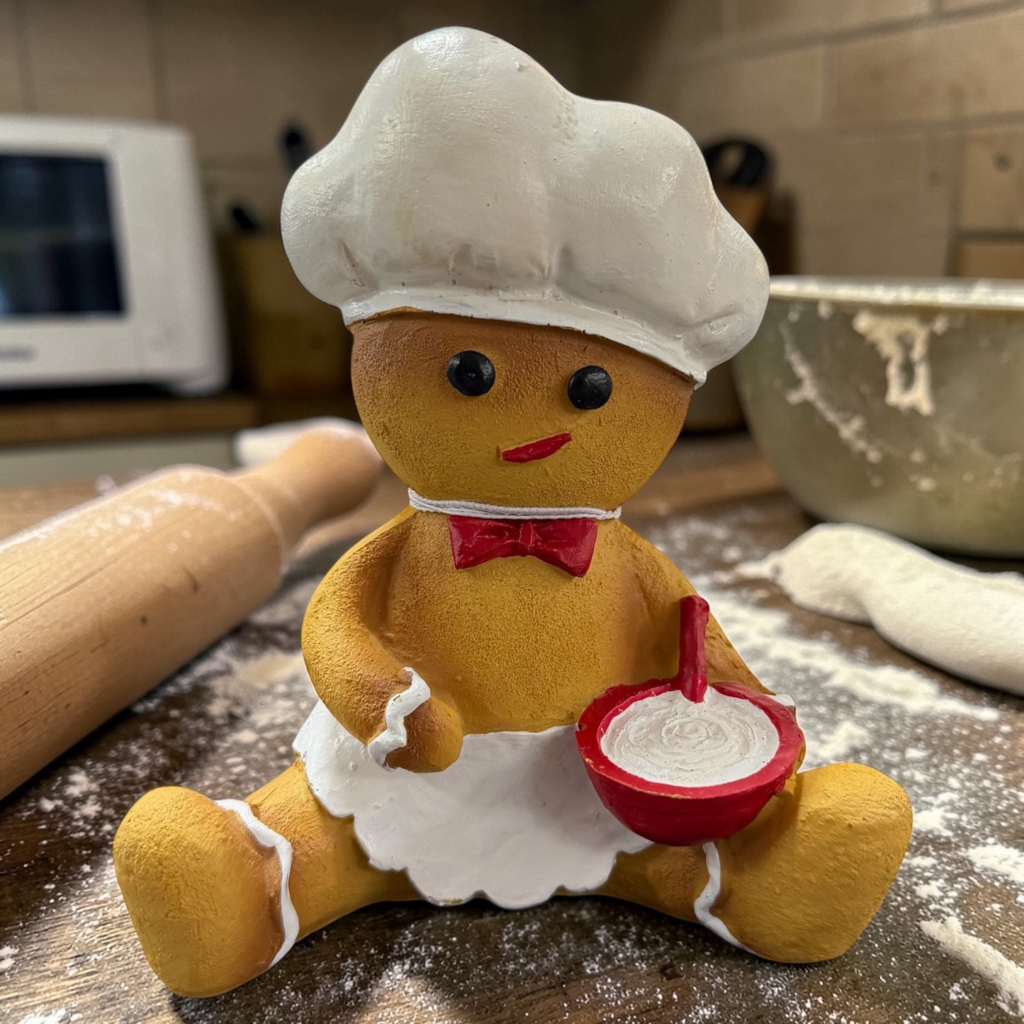 Eine Dekofigur in Form einer Lebkuchenfigur in Braun mit weißer Schürze und roter Schleife sowie roter Schüssel, platziert auf einer bemehlten Arbeitsfläche mit Nudelholz und Schüssel im Hintergrund.