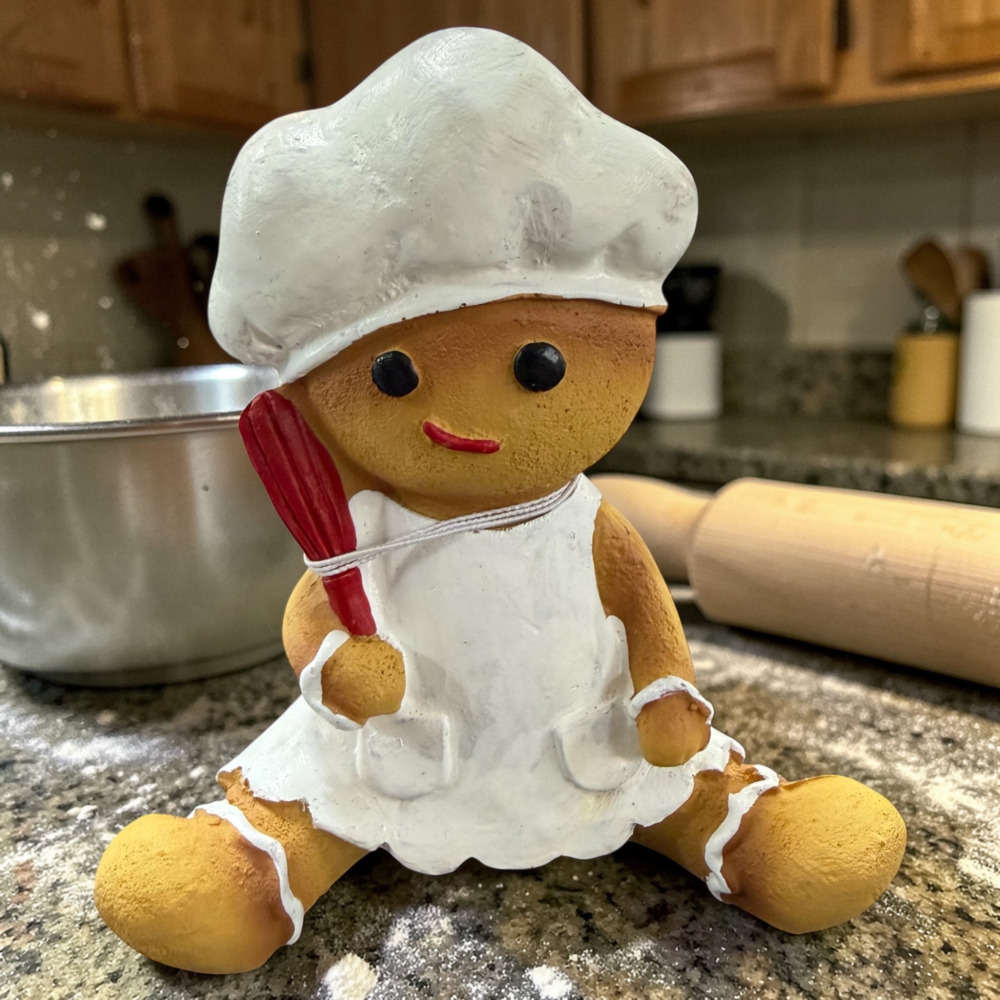 Eine Dekofigur in Form einer Lebkuchenfigur in Braun mit weißer Kochmütze und Schürze sowie rotem Schneebesen, dargestellt auf einer bemehlten Arbeitsfläche mit Schüssel und Nudelholz im Hintergrund.
