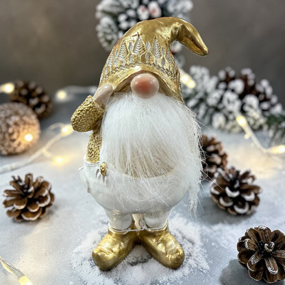 Ein Wichtel mit goldener Mütze, weißem Bart und erhobener Hand steht zwischen Tannenzweigen, Zapfen und Lichterkette in winterlicher Dekoration.