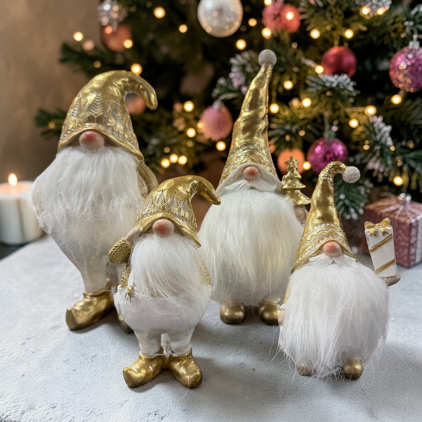 Vier Wichtel in verschiedenen Größen mit goldenen Details und weißen Bärten stehen dekorativ vor einem geschmückten Weihnachtsbaum mit Lichterkette.