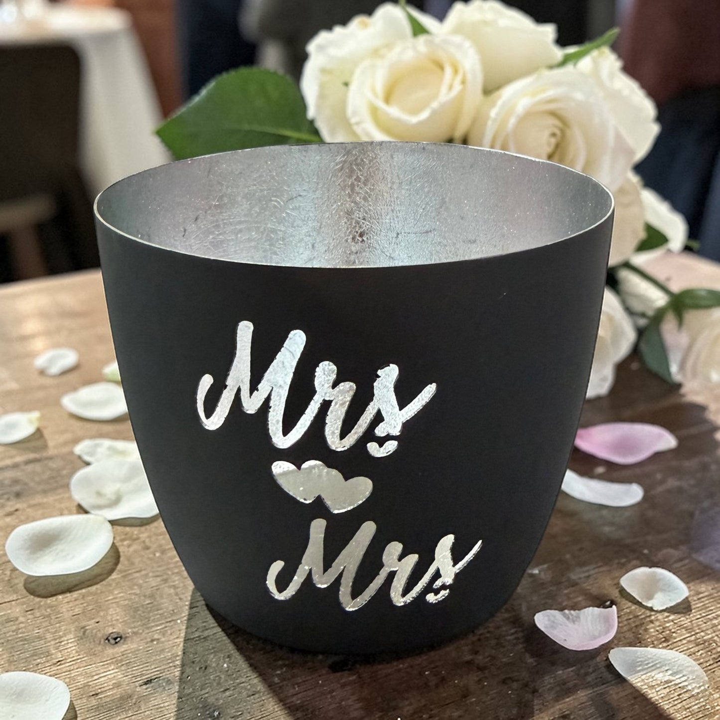 Ein Windlicht aus Metall in Schwarz mit silberner Innenfläche, dargestellt auf einem Holztisch mit Blumen und Hintergrundbeleuchtung, mit dem Schriftzug „Mrs & Mrs“.