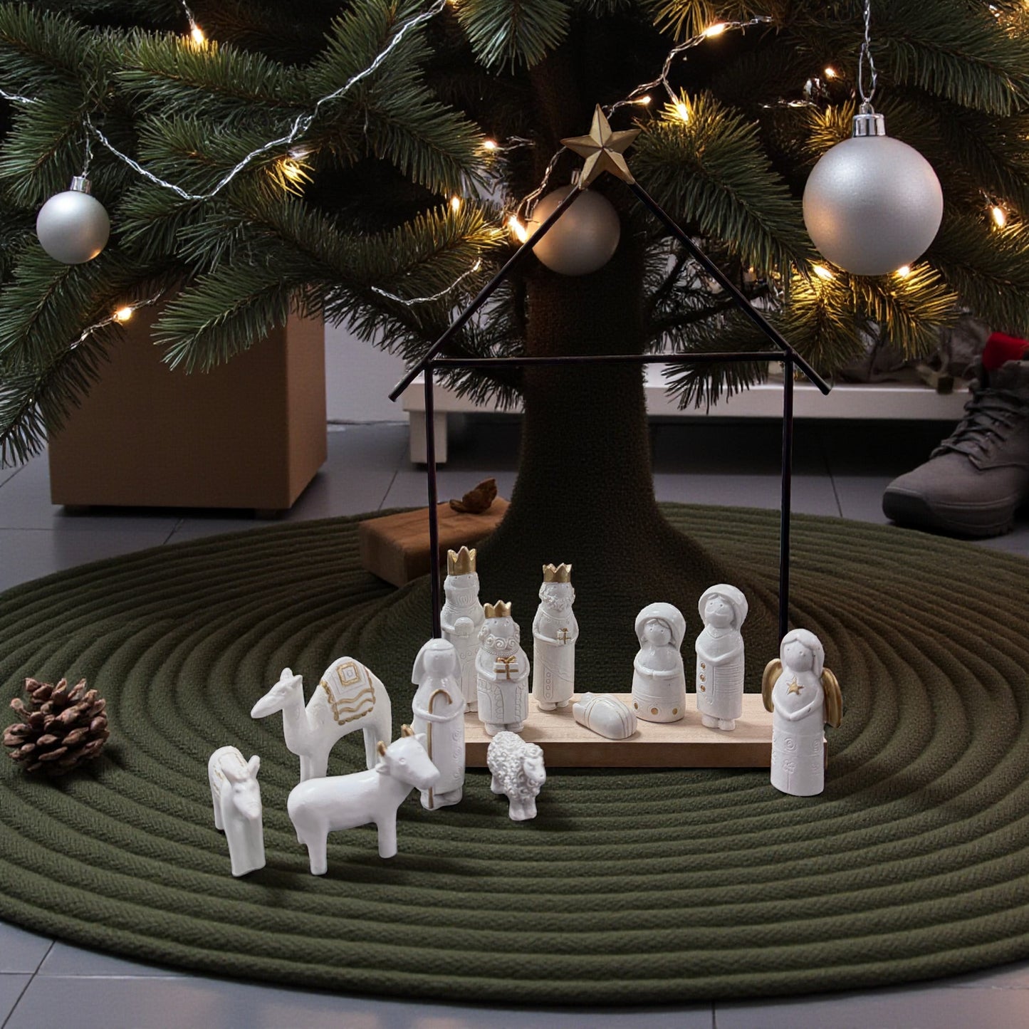 Moderne Weihnachtskrippe mit weißen Keramikfiguren und goldenen Details in schwarzem Metallgestell in Hausform, dekoriert unter Weihnachtsbaum mit Lichterkette und Kugeln, platziert auf grünem Teppich.