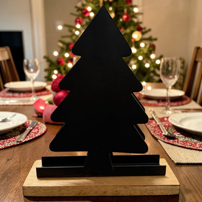 Ein schwarzer Serviettenhalter in Tannenbaumform steht auf einem Holzsockel auf einem gedeckten Tisch, im Hintergrund ist ein beleuchteter Weihnachtsbaum zu sehen.