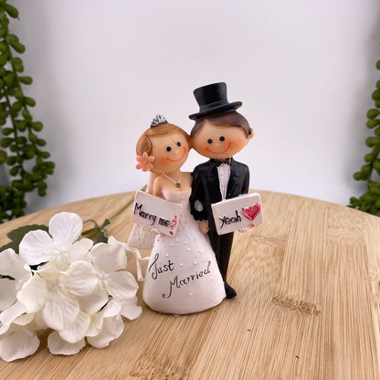 Hochzeit Dekofigur Tortenaufsteller im Ambiente bildet Brautpaar Mann & Frau ab. Das Ehepaar hält sich an der Hand. Die Frau hält ein Schild mit "Marry Me?" und der Mann mit "Yeah". Auf dem weißen Brautkleid steht Just Married. 