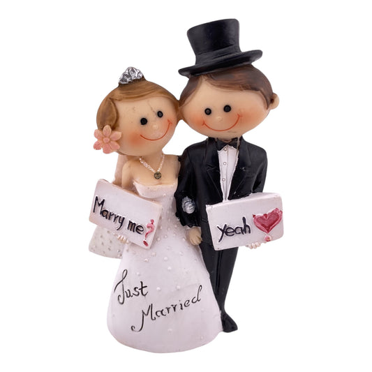 Hochzeit Dekofigur Tortenaufsteller vor weißem Hintergrund bildet Brautpaar Mann & Frau ab. Das Ehepaar hält sich an der Hand. Die Frau hält ein Schild mit "Marry Me?" und der Mann mit "Yeah". Auf dem weißen Brautkleid steht Just Married.