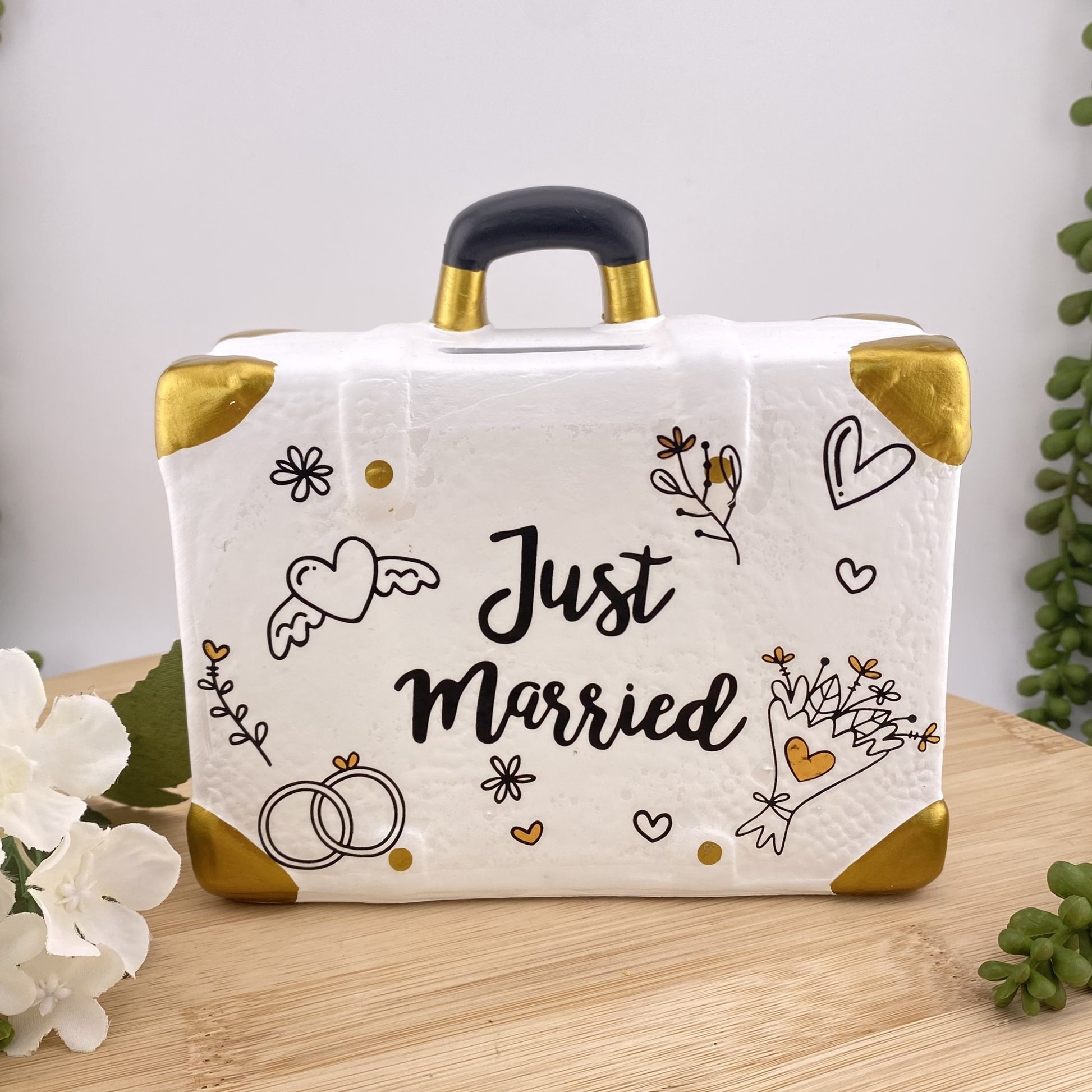 Eine weiße Spardose in Form eines Koffers mit goldenen und schwarzen Symbolen und dem Schriftzug Just Married in der Mitte. Das Hochzeitsgeschenk ist im Ambiente  platziert.