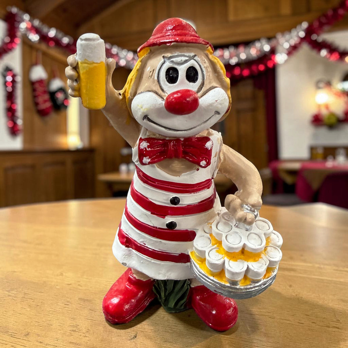 Karneval Deko Clown Die Schmitzens Köbes mit Kölschglas und Tablett vor rot-weißer Karnevalsdekoration.