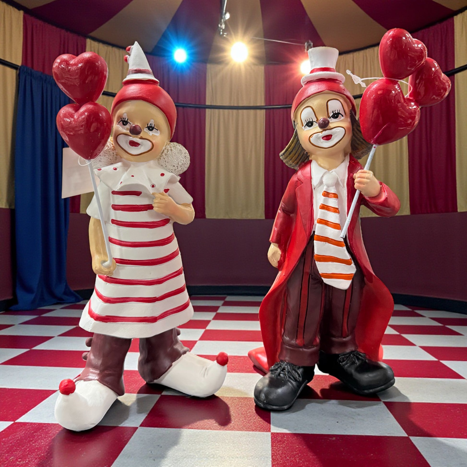 Zwei dekorative Clownfiguren mit roten Mänteln und rot-weiß gestreifter Kleidung, jeweils mit herzförmigen Ballons, stehend in einer farbig beleuchteten Zirkus- bzw. Karnevalskulisse.