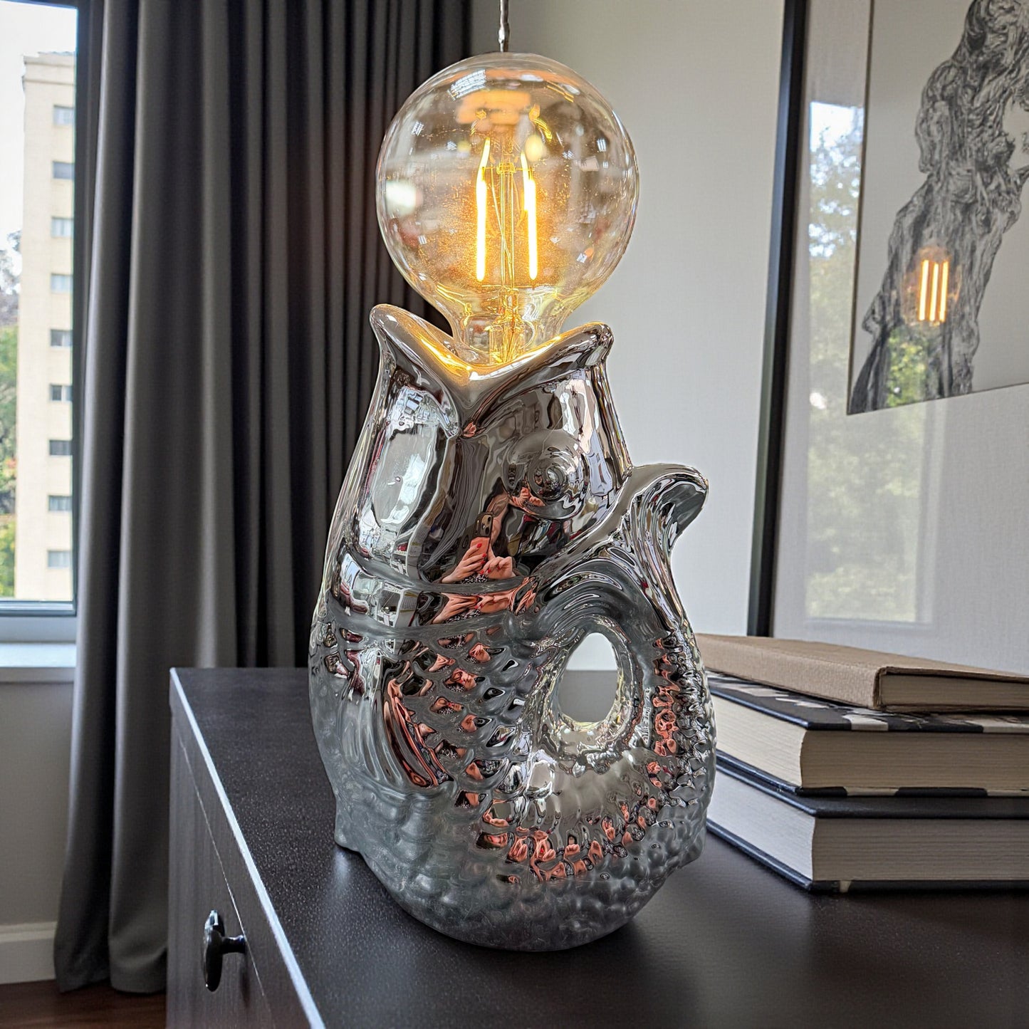 Dekorative Keramiklampe in Fischdesign mit glänzender Oberfläche und warm leuchtender Filament-Glühbirne, platziert auf Sideboard neben Büchern im Wohnraum-Ambiente mit Fenster und Vorhängen.