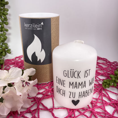 Eine kleine, weiße Kerze mit schwarzem Schriftzug "Glück ist eine Mama wie dich zu haben". Die Dekokerze ist 8cm groß und im Ambiente platziert.