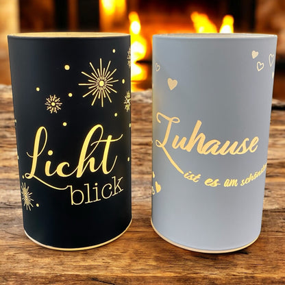 Zwei LED-Deko-Lichtsäulen in Schwarz und Weiß mit goldenen Sprüchen „Lichtblick“ und „Zuhause ist es am schönsten“ im Ambiente
