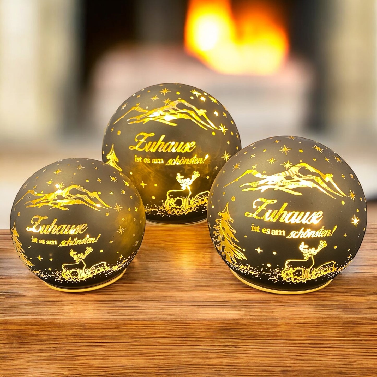 Leuchtende goldene Dekokugeln mit Weihnachtsmotiv und Spruch „Zuhause ist es am schönsten“ vor Kaminhintergrund.