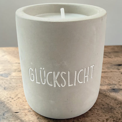 Graue Betonkerze mit eingraviertem Schriftzug „GLÜCKSLICHT“ auf einer Holzoberfläche.