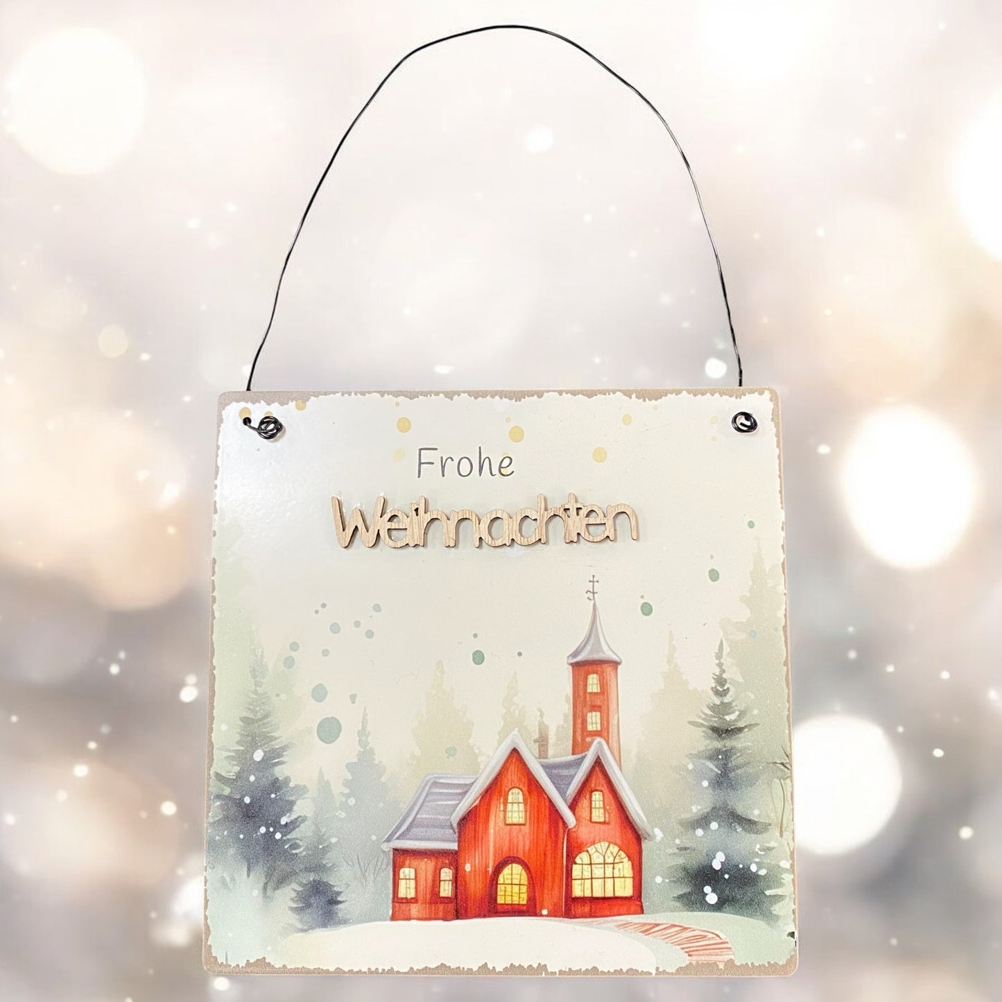 Metallschild mit Aufschrift „Frohe Weihnachten“ und Motiv einer roten Kirche im verschneiten Wald, vor hellem Bokeh-Hintergrund.

