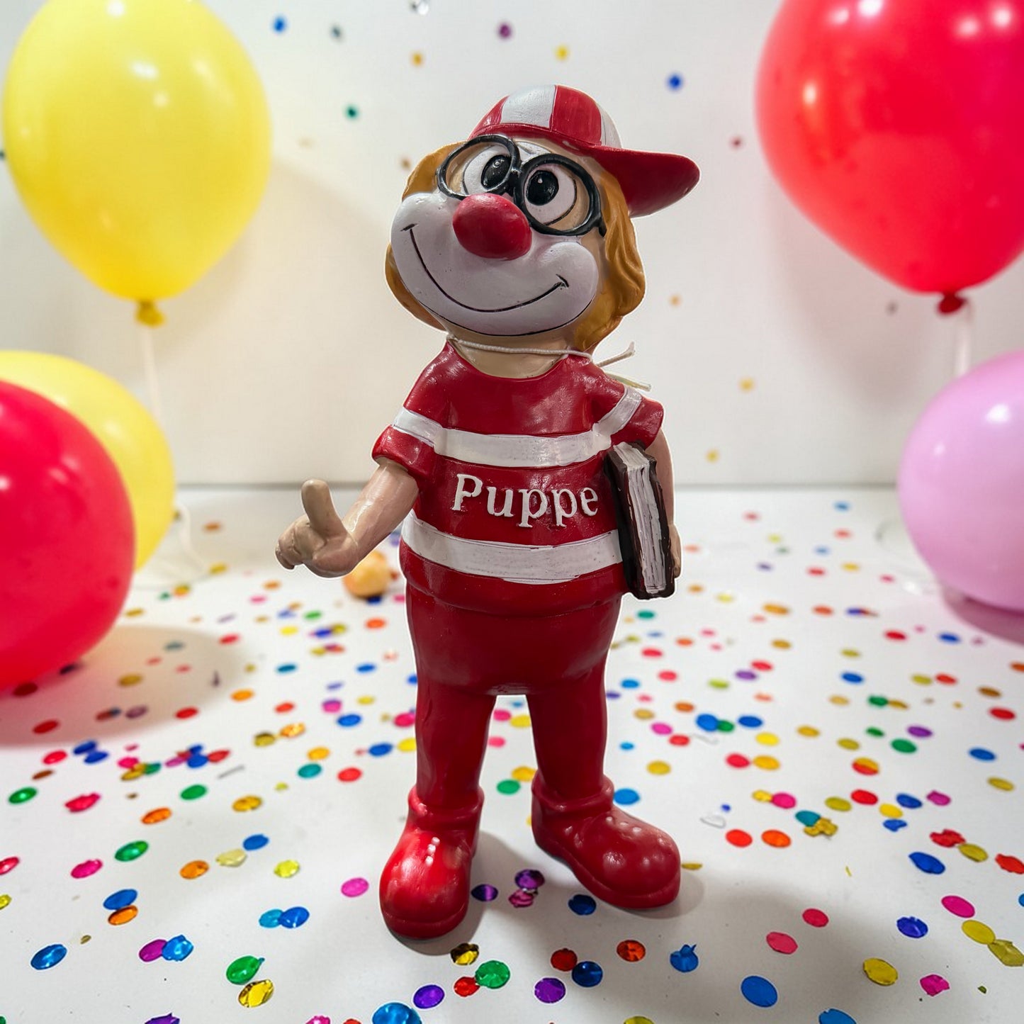 Clown-Dekofigur in Rot mit Kappe, Brille und Buch unter dem Arm vor bunten Luftballons und Konfetti.