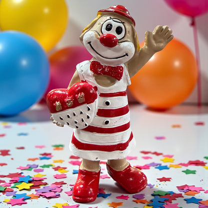 Clown-Figur mit roter Nase und rot-weiß gestreiftem Kleid, eine Hand erhoben und ein herzförmiges Objekt haltend, stehend auf einer Tischfläche mit Konfetti, im Hintergrund farbige Luftballons.