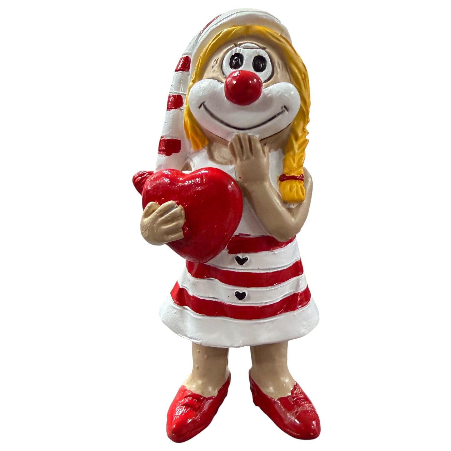 Clown-Figur in rot-weißem Kleid mit roter Nase, hält ein rotes Herz vor dem Körper und eine Hand am Gesicht, vor weißem Hintergrund.
