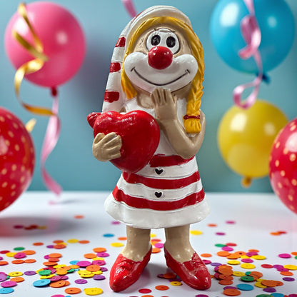 Clown-Figur in rot-weißem Kleid mit roter Nase, hält ein rotes Herz vor dem Körper und eine Hand am Gesicht, stehend auf einer Tischfläche mit Konfetti, im Hintergrund farbige Luftballons.