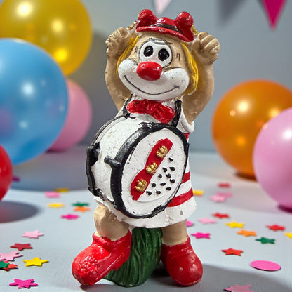 Clown-Figur mit roter Nase und Trommel vor dem Körper, die Arme erhoben, stehend auf einer Tischfläche mit Konfetti, im Hintergrund farbige Luftballons