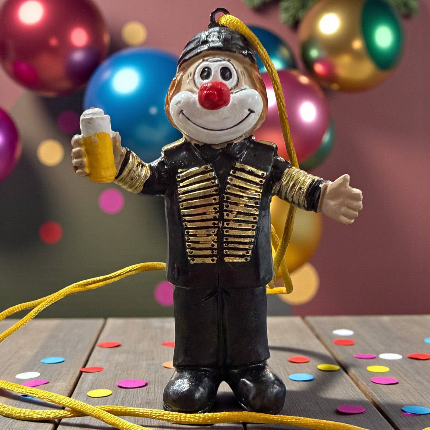 Clown-Figur mit roter Nase in schwarzer Uniform mit goldfarbenen Details, hält einen Bierkrug in einer Hand, mit Aufhängeband, stehend auf einer Holzfläche mit Konfetti, im Hintergrund bunte Kugeln.