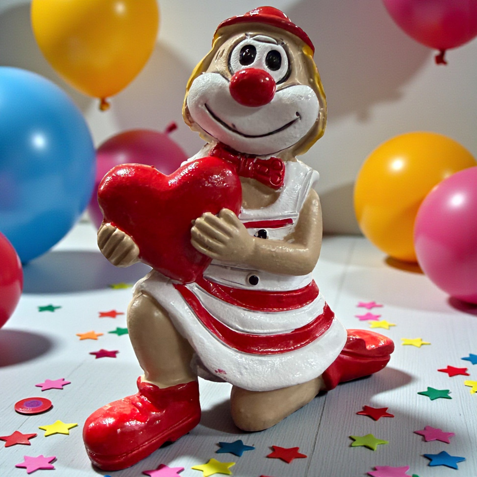 Clown-Figur mit roter Nase in rot-weißem Outfit, kniend dargestellt und ein rotes Herz haltend, auf einer Tischfläche mit Konfetti, im Hintergrund farbige Luftballons.